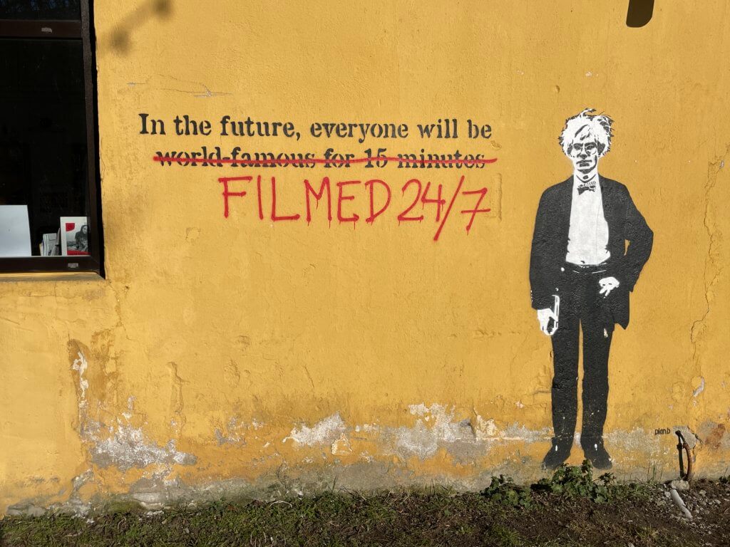 A graffitied wall showing Andy Warhol with the text, "In the future, everyone will be world famous for 15 minutes." The text "world famous for 15 minutes" has been crossed out and "Filmed 24/7" has been spray painted below. 